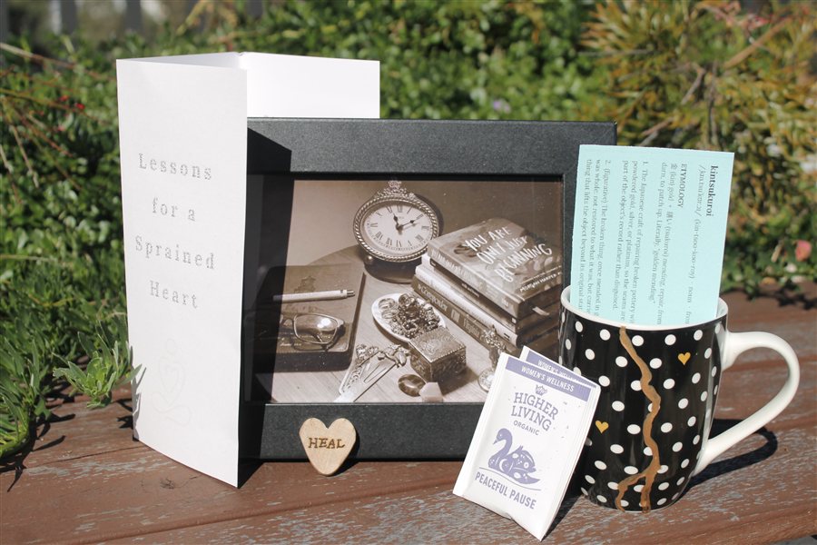 Golden Mending flat lay — a folded poem card, framed sepia photograph, wooden heart and mug of tea